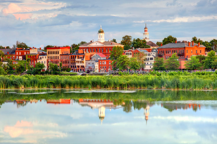 Springtime view of Exeter, New Hampshire, from across the Squamscott River. Thick green grasses grow along the banks of the river, between the water and the city’s buildings.