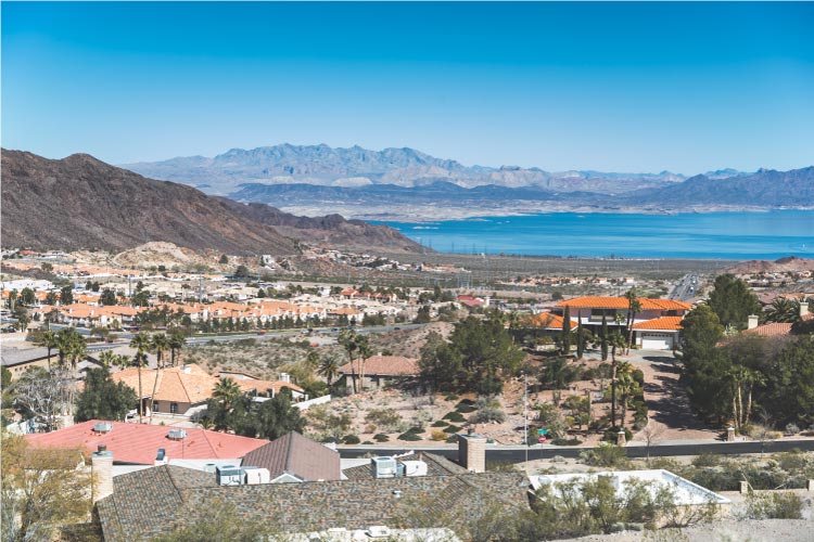 Rooftop view of Boulder City, Nevada. The town is built into a desert landscape, with palm trees and other vegetation dotting the spaces between houses. Lake Mead and surrounding mountains are visible in the distance.
