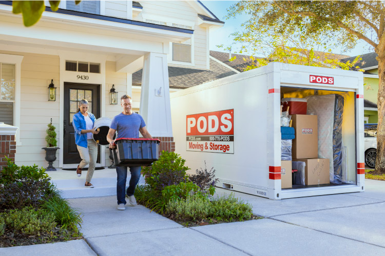 A mature couple is carrying a plastic tub and footstool out to the PODS portable moving container in their driveway. The container is already loaded with a mattress, moving boxes, and other furniture pieces. 