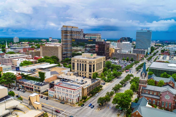 Aerial view of downtown Columbia, South Carolina. There’s a mix of tall and squat city buildings, as well as clusters of mature trees growing along streets and between city blocks. 