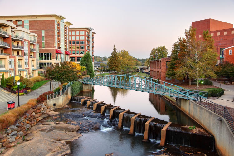 View of the downtown part of Greenville, South Carolina. A pedestrian bridge crosses a river above a small dam. Large red brick buildings line the river on either side. 