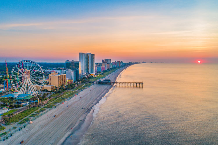 Aerial view of Myrtle Beach, South Carolina, during sunset. Along the white sand beach are tall condo buildings, a ferris wheel, and a pier.