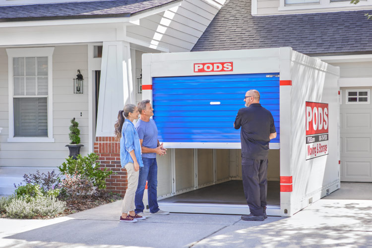 A senior couple is speaking with a PODS truck driver about their PODS portable moving container. The driver is opening the blue door of the container to show them the inside. 