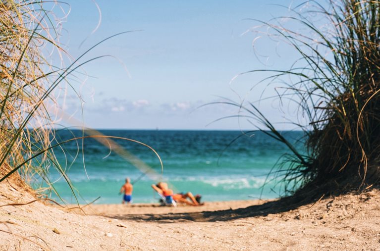 The view through a sandy dune path reveals a mature couple enjoying a relaxing day at the beach after retiring in Florida. The man is fishing, and the woman is sunning nearby.