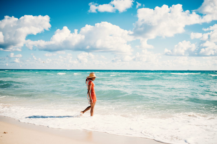 A woman is wearing an orange sun dress and a straw hat as she walks through the gentle surf of Miami Beach. 