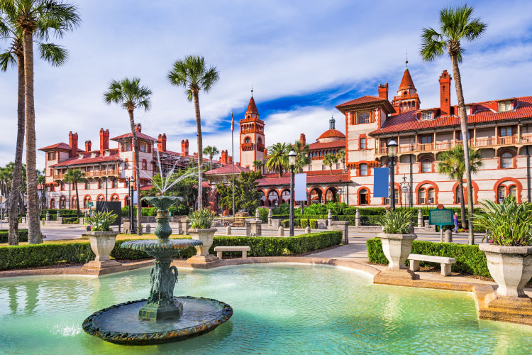 A view of downtown buildings in St. Augustine, Florida. The buildings have a Spanish flair to them, with light exteriors and bright red tile roofs. In the foreground is a large, decorative fountain. 