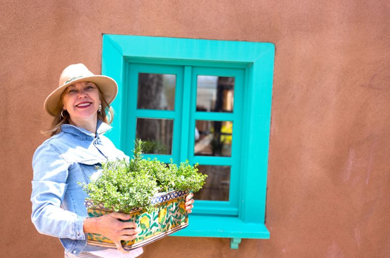 A senior woman is smiling in front of a classic New Mexico adobe house, holding an ornate flower box.