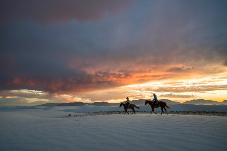 Two people ride horses through White Sands National Park in New Mexico during sunset. Low-lying clouds reflect the last rays of sunlight, casting a magical glow across the sky, and mountain ranges dot the horizon.