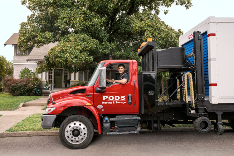 A PODS truck driver is smiling from the cab of his truck, parked in front of a residential home. There’s a PODS portable moving and storage container on the back of the truck. 