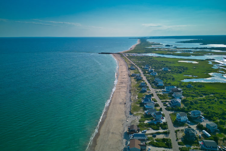 Aerial view of the pristine beach in Charlestown, Rhode Island, on a clear summer day. Beach houses dot the shore, and a small inlet in the distance creates a path for ocean water to make its way inland.