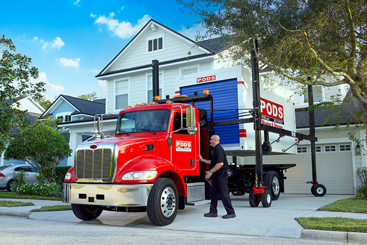 A PODS truck is backed into a residential driveway. There’s a PODS portable moving container on the bed of the truck, and the driver is preparing to use PODZILLA to position the container in the driveway so it can be loaded.