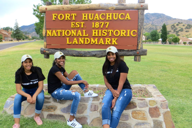 Three women smile for a photo in front of the Fort Huachuca Historic Landmark sign at Fort Huachuca in Sierra Vista, Arizona.