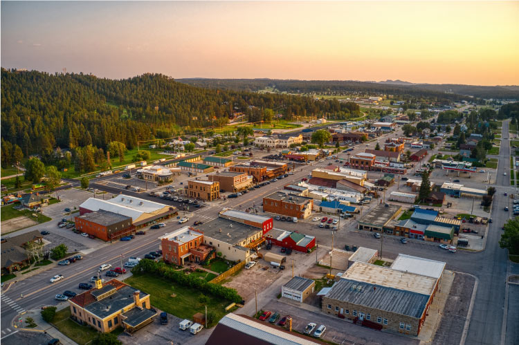Aerial view of the small town of Custer, South Dakota, in the summer. The hills on the outskirts of the town are covered in lush pine trees, and the city buildings are spaced wide apart.