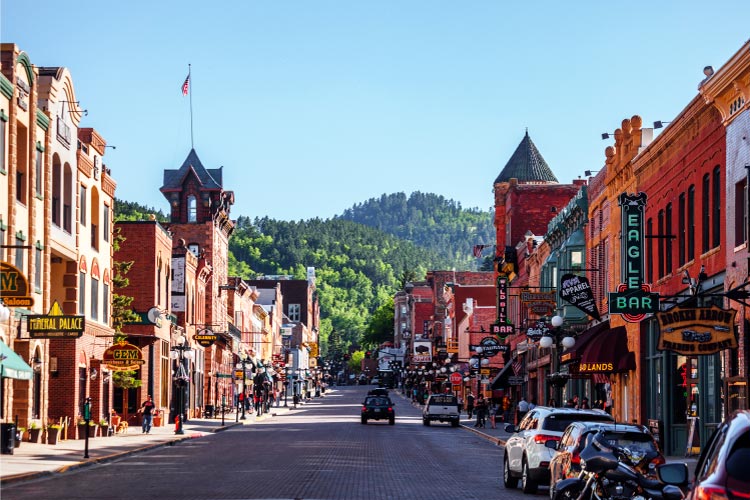 The view down a historic street in Deadwood, South Dakota, on a sunny summer day. Most of the buildings are made of red brick and a lush tree-covered hill rises up outside of town.