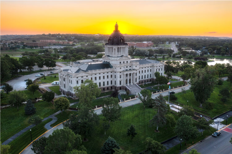 Aerial view of the state capitol building in Pierre, South Dakota, during sunset. The lush green of surrounding green spaces and trees contrasts beautifully with the bright yellow and orange sky.