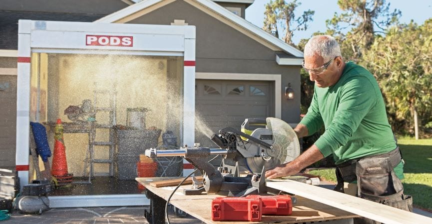 A mature man is cutting a board of wood on a saw table in his driveway. Behind him is an open PODS storage container, which is being used to store construction supplies during a home renovation project.