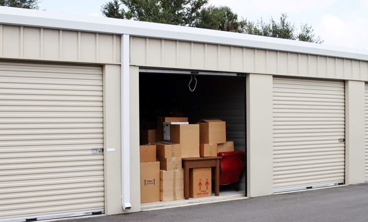 Exterior view of several side-by-side outdoor self-storage units that are painted beige. The center unit has its door open. Inside is some furniture and many cardboard boxes. 