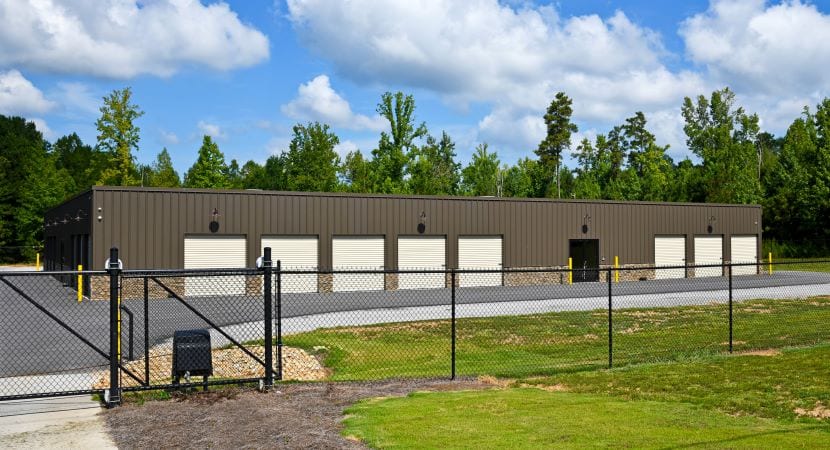 A secure storage center with a chainlink fence surrounding it. The building is dark brown, and each storage unit has a light-colored garage-style door.
