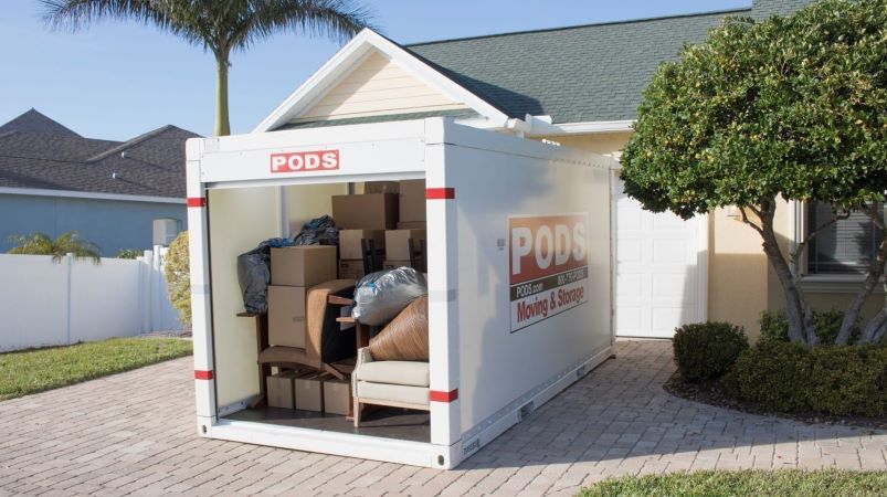  A PODS portable moving and storage container is sitting in a residential driveway in Florida. It is 75 percent loaded with moving boxes and furniture, but there is still room left for more. The door of the container is open, showing what’s inside. 