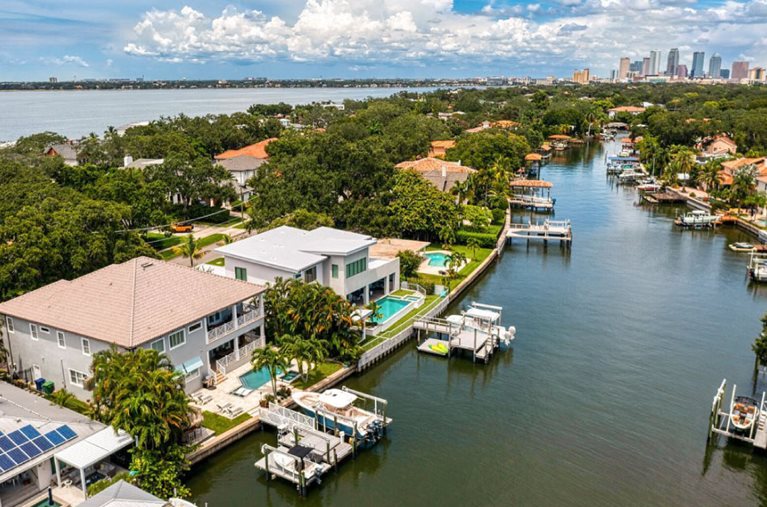Aerial view of the Tampa neighborhood Davis Island. Luxury waterfront mansions dot the waterway, each with its own private dock. In the distance, Tampa’s skyline can be seen against a cloudy blue sky.
