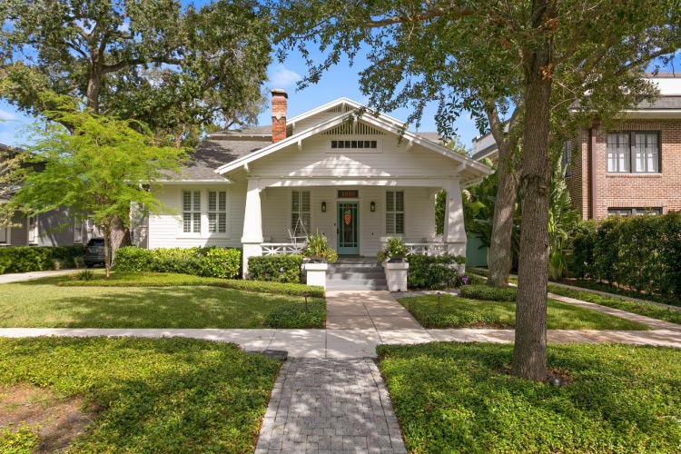A lovely white bungalow in Tampa neighborhood historic Hyde Park. The home features a wide covered porch, a chimney, and a shaded lawn.