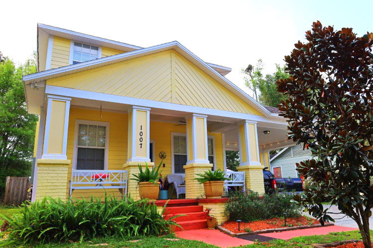 A cute yellow house in Tampa’s Old Seminole Heights neighborhood features a large covered porch with boxy columns and a gable roof.