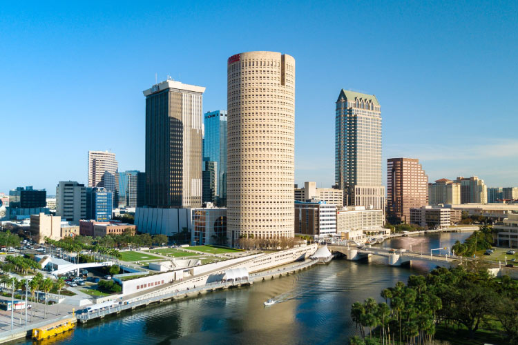 View of Downtown Tampa from across the river on a sunny day. Several distinct buildings make up the skyline, and a boat makes its way along the river.
