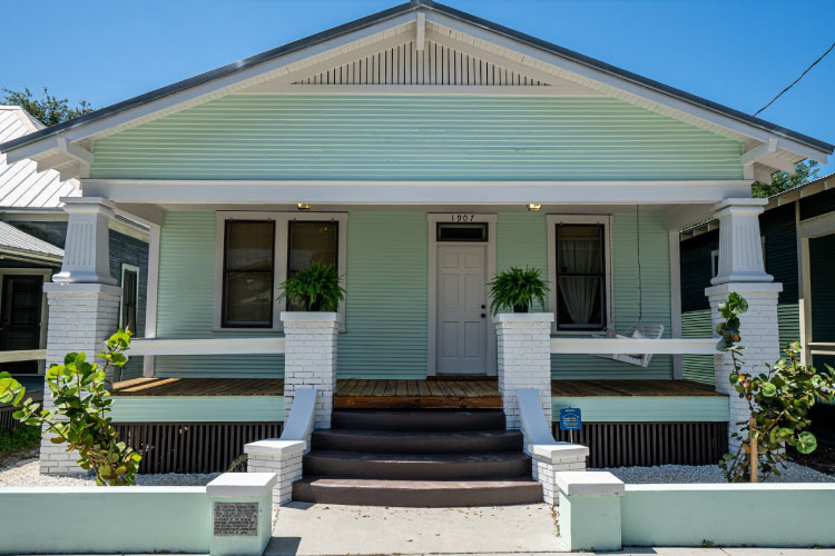 A white and light green bungalow in Ybor City with a large covered porch and seashell yard. A couple of native plants decorate the yard.