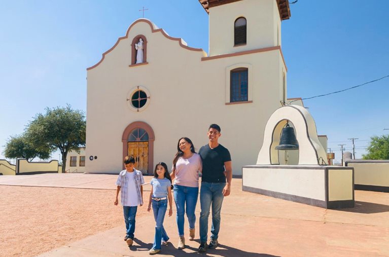 A family of four is walking in front of an adobe church in El Paso, Texas, on a sunny day.