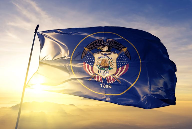 The state flag of Utah, blowing in the wind at the top of a flag pole as the sun sets behind a range of mountains in the background