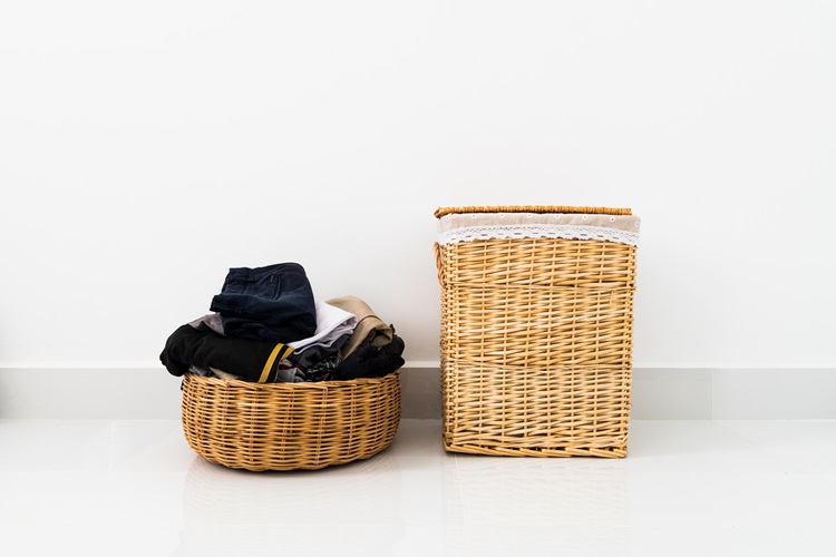 Two baskets sitting side-by-side on the floor. The one on the right is tall and has a lid, and the one on the left is short and round with an open top. The basket on the right is meant to be used for clothing that should be thrown away, and the one on the left is filled with clothing to be donated. These baskets can be kept in a closet to help keep it from becoming cluttered.