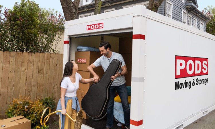 A couple is in the process of moving things into a PODS portable storage container to help them declutter. The woman is handing a guitar case to the man and they’re both smiling. The container already has many packed boxes in it.