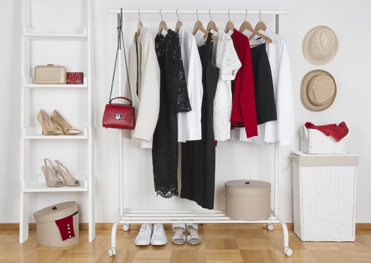 A well-organized closet clothes rack holds hanging clothing and a purse. There’s a shelf to the left of the rack, with shoes and a couple of clutch purses. On the other side is a basket with two hats hanging on the wall above it.
