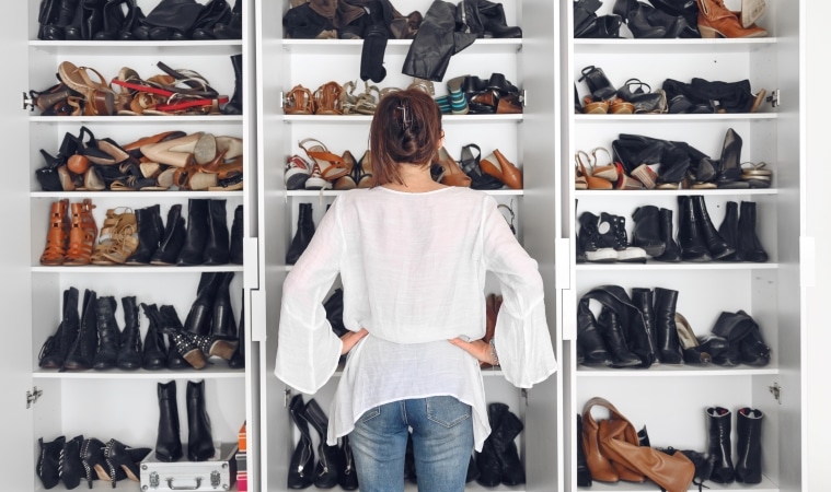 A woman is standing, facing her shoe closet with her back to the camera. The shelves of the closet are overflowing with dozens of pairs of shoes and boots. 