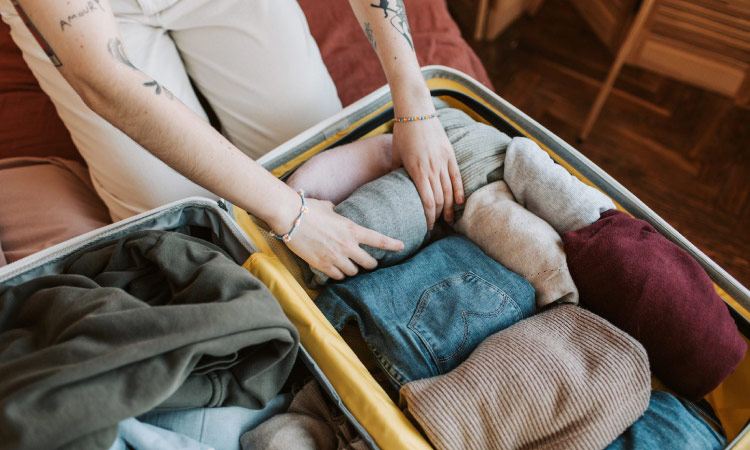 A person with tattoos on their forearms is packing cool-weather clothing in a suitcase for storage. The suitcase is full of sweaters, jeans, and sweatshirts.