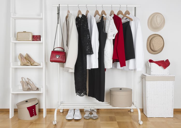 A well-organized clothes rack has clothes and a purse on it. There’s a shelf beside it on one side with shoes and a handbag. On the other side is a basket with two hats hanging above it.