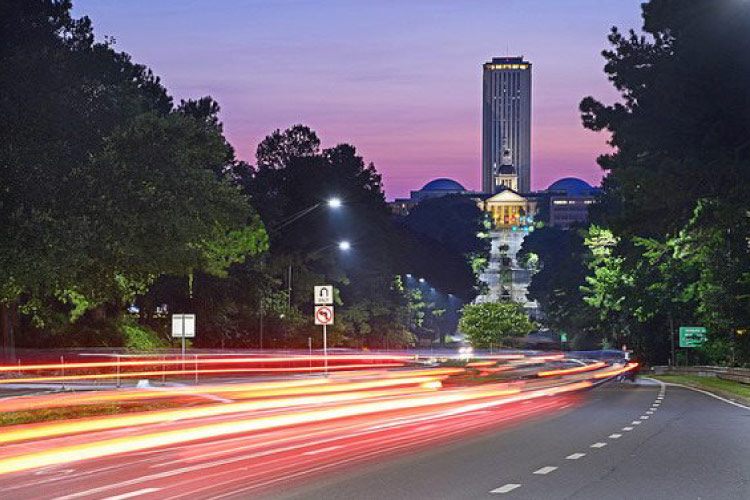 Traffic zips along Tallahassee’s highway US-27 at dusk with the city’s capitol in the background. The cars are all a blur of red and yellow lights. 