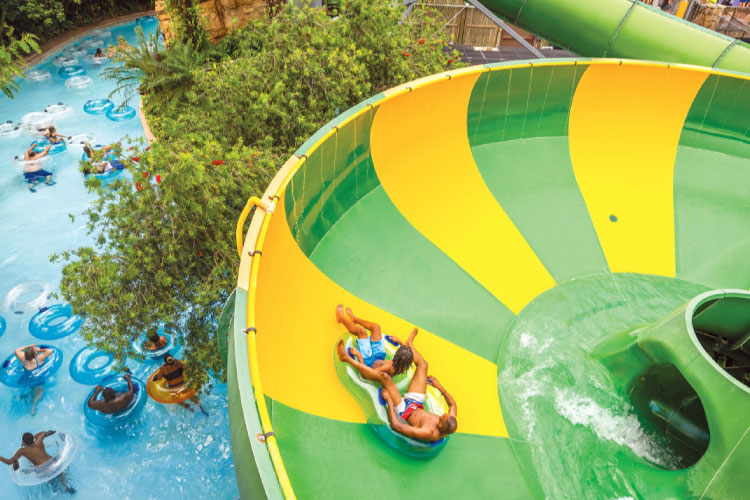 Two friends ride a tandem figure-eight tube down a green and yellow water slide at the Adventure Island water park in Tampa, Florida. Below the water slide, dozens of people enjoy a leisurely float around a lazy river.