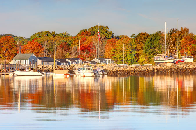 Quaint buildings, moored and beached sailboats, and vibrant autumn foliage reflect in the tranquil waters along a rocky shore in Rye — one of the best towns in New Hampshire.
