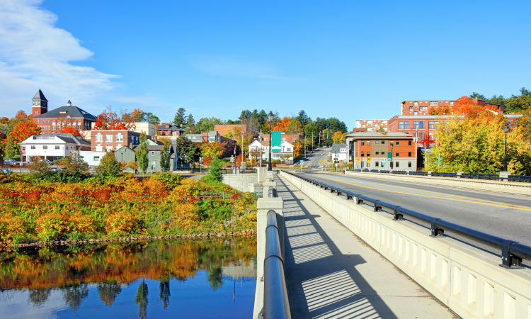 View of the charming town of Plymouth, New Hampshire, captured from across a sturdy bridge. The scene is set in late summer, as the foliage begins its gradual transformation, showcasing trees with hints of changing colors. Bathed in abundant sunlight, the brick buildings of the town exude a vibrant radiance against the backdrop of the blue summer sky.