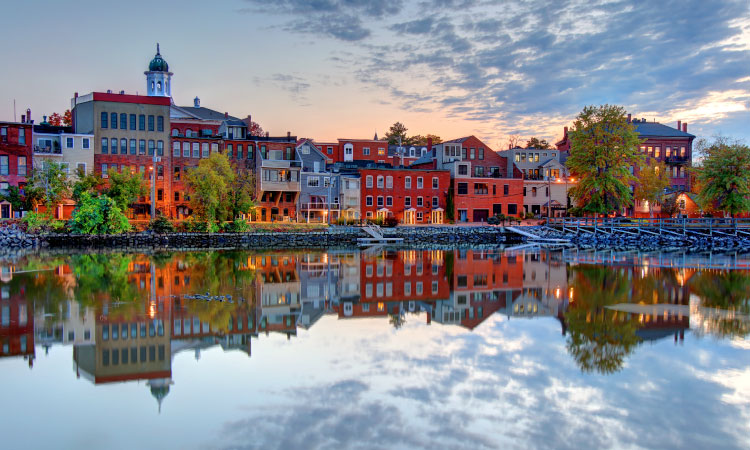 A captivating twilight scene showcasing Exeter, New Hampshire, viewed from across the tranquil Squamscott River. The town's mostly red brick buildings stand prominently, their charming architectural details illuminated by the soft glow of the setting sun. The serene river mirrors the town's image, creating a captivating visual symmetry.