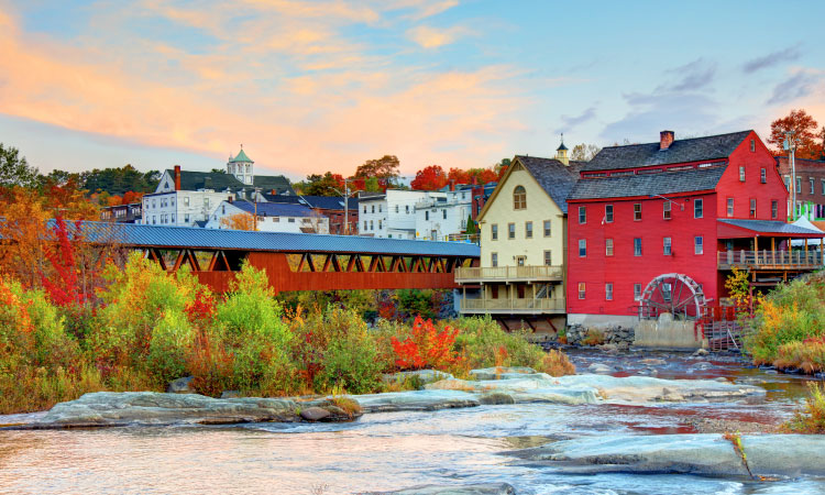 A scenic view of the picturesque town of Littleton, New Hampshire, framed by the iconic covered bridge as observed from across the flowing Ammonoosuc River. The riverbank is adorned with historic structures, notably the charming red brick Renaissance Mills of Littleton, adding to the town's timeless allure.