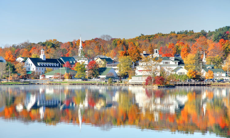 A scenic view of Meredith, New Hampshire, as observed from across the serene Lake Winnipesaukee. The autumnal ambiance is evident, with trees displaying a mix of vibrant oranges and reds, some already bare. The tranquil lake mirrors the town's numerous white structures, including charming churches with prominent steeples, creating a picturesque reflection on its still surface.