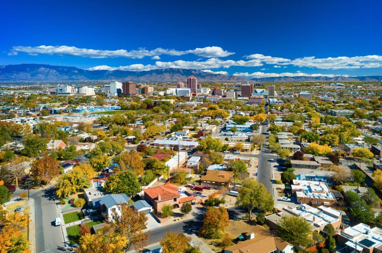 Aerial view of Albuquerque, New Mexico, and its surrounding neighborhoods and residential areas. It’s late summer and some of the trees have started to change colors. The sky is a vibrant blue and the Sandia Mountain Range is visible in the distance.