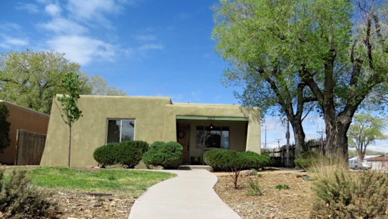 Exterior view of a one-story adobe bungalow in the Nob Hill neighborhood of Albuquerque, New Mexico, featuring pruned bushes by the entrance.