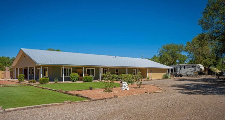 A large, one-story ranch home in the South Valley neighborhood of Albuquerque, New Mexico. The home is surrounded by a wraparound awning and a simple, green lawn. There’s a travel camper parked behind the home.