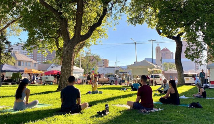 Albuquerque locals practice yoga in the park in Downtown Albuquerque. Seven students are sitting on yoga mats in the park’s green field, under the shade of tall trees. In the distance, market tents are set up for an event and downtown buildings are illuminated in the mid-morning light.