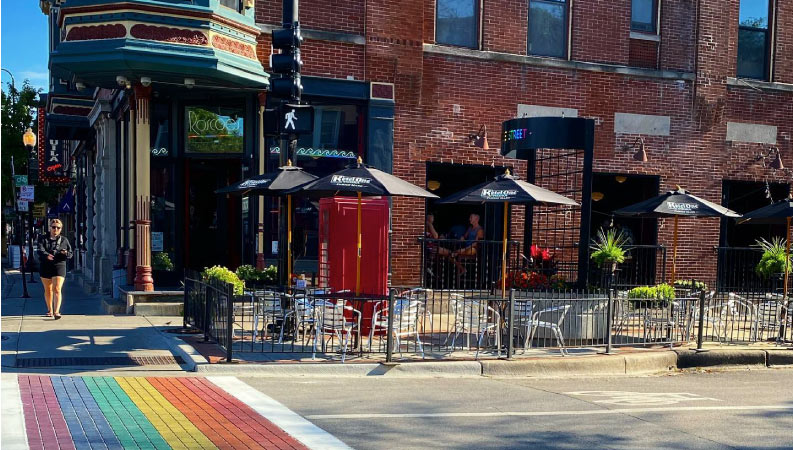 A woman is about to walk across a rainbow-painted crosswalk outside Roscoe’s Tavern in the Northalsted neighborhood of Chicago, Illinois. It’s a sunny day, so she’s wearing shorts and sunglasses.