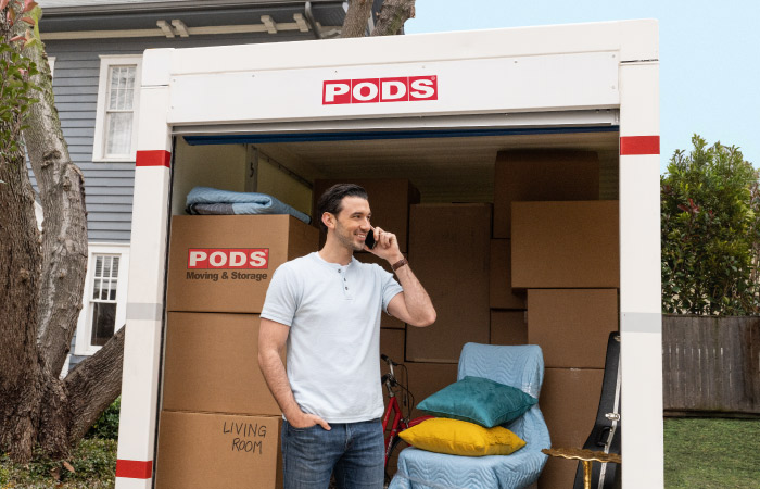A man is talking on his cell phone at the entrance to his PODS portable moving container. The container is almost fully loaded with moving boxes, furniture, and other items.