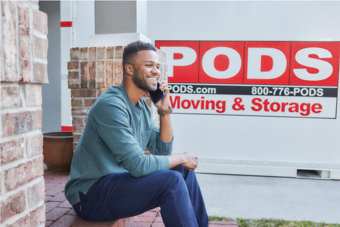 A young man is making a phone call on his front step beside the PODS portable moving container in his driveway